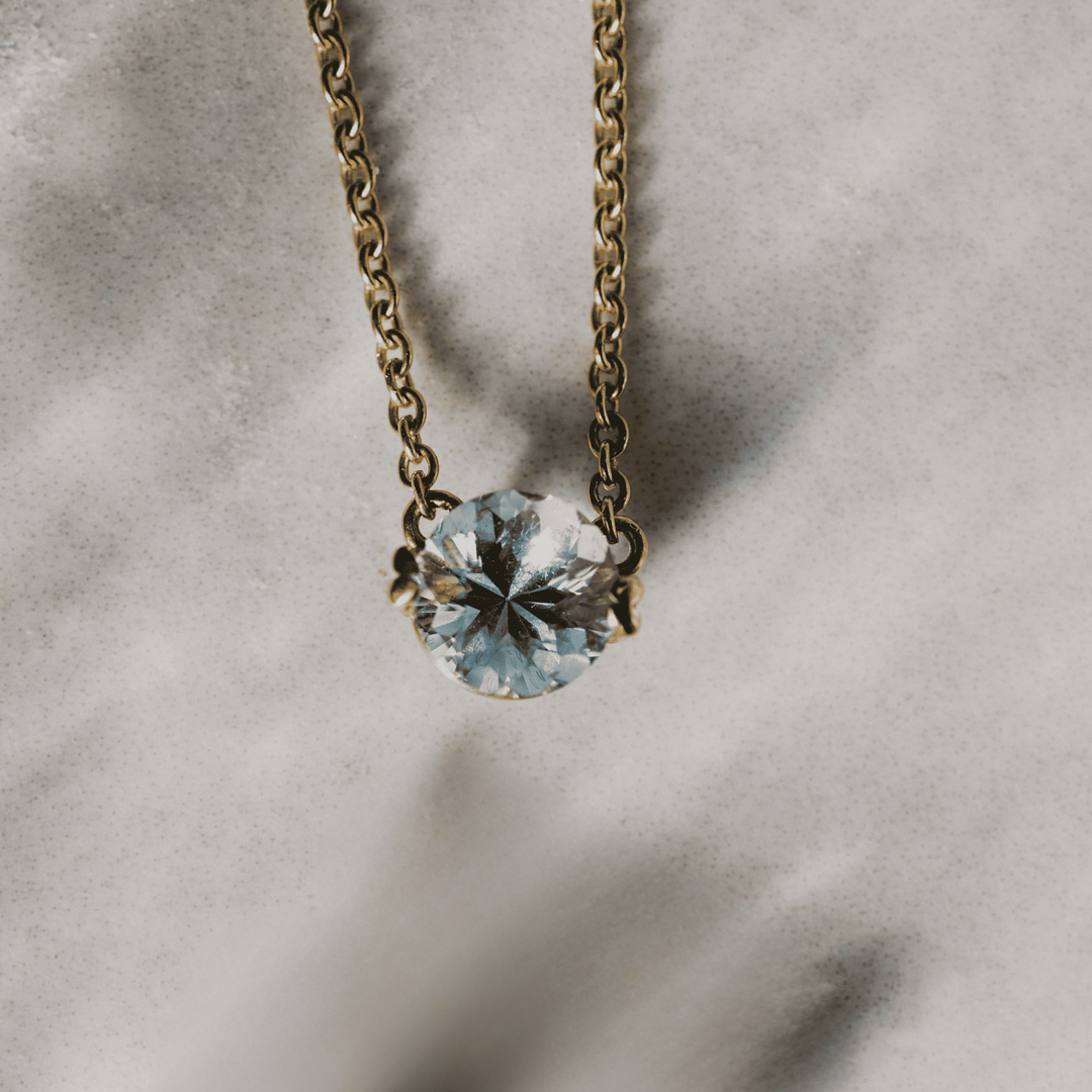 Gelbgoldkette mit rundem, facettiertem Aquamarin-Anhänger vor sandigem Hintergrund