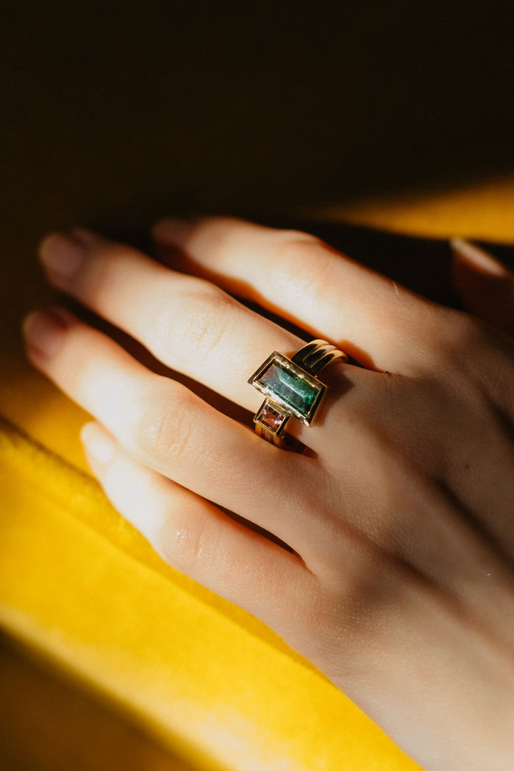 Hand mit dem Mon Trésor Ring aus Gelbgold und grünem Turmalin, fotografiert im warmen Sonnenlicht auf gelbem Samt.
