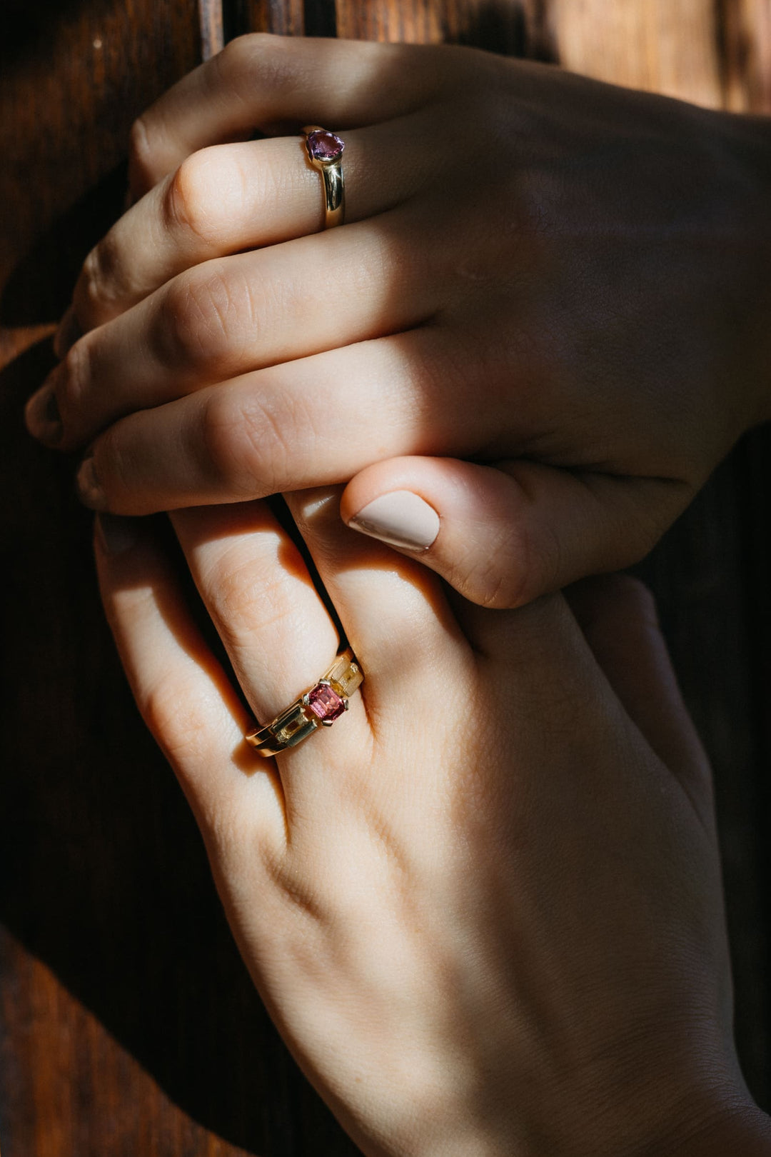 Zwei übereinandergelegte Hände mit dem Le Pont Ring aus Gelbgold und pinkem Turmalin vor einer hölzernen Struktur