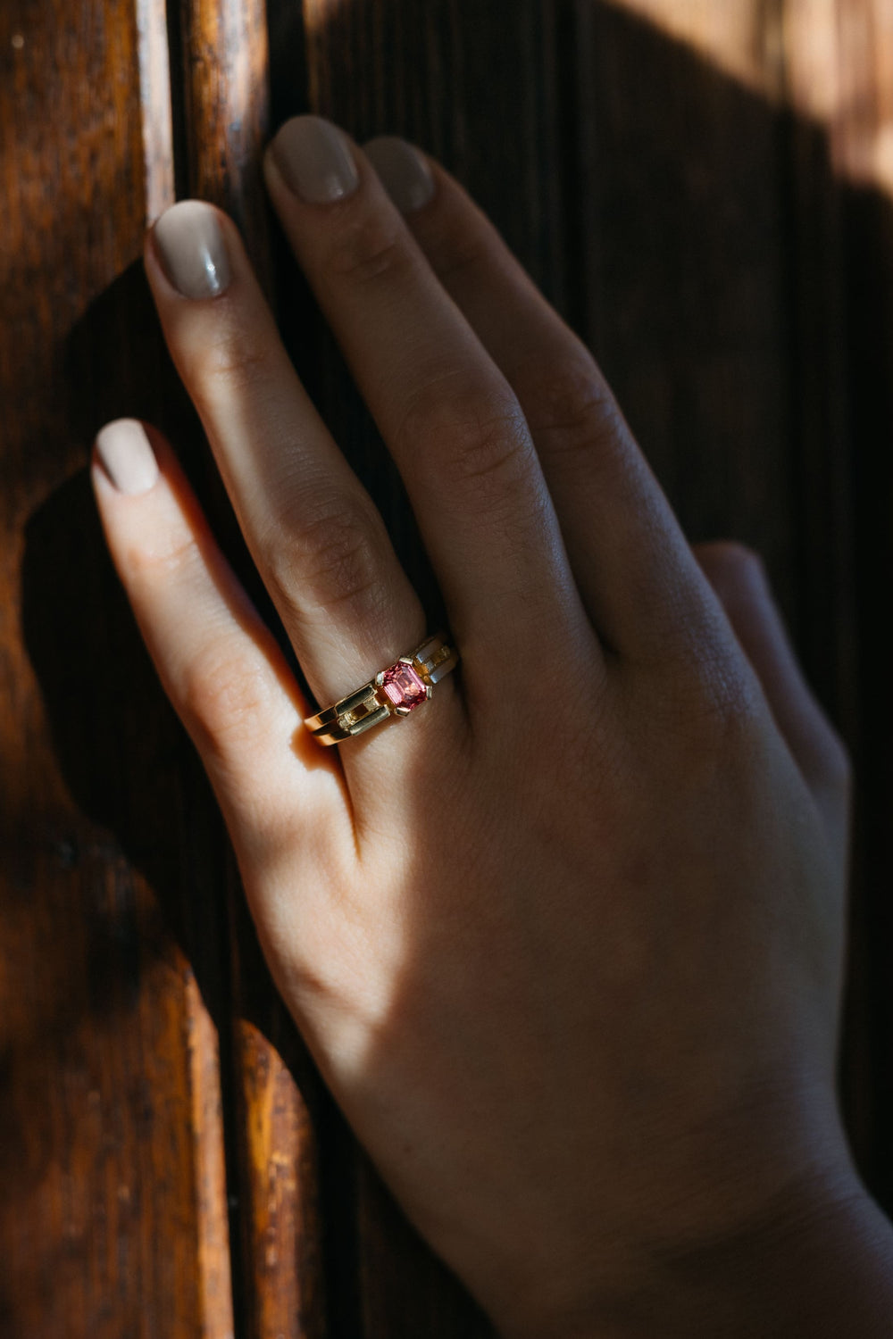 Hand mit dem Le Pont Ring aus Gelbgold und pinkem Turmalin vor einer hölzernen Oberfläche im Sonnenlicht