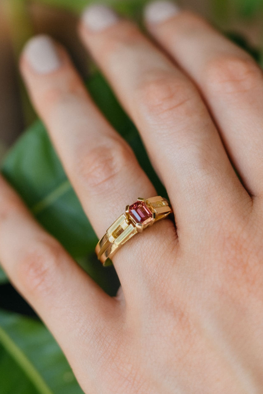 Hand mit dem Le Pont Ring aus Gelbgold und pinkem Turmalin, gehalten über grünen Pflanzenblättern