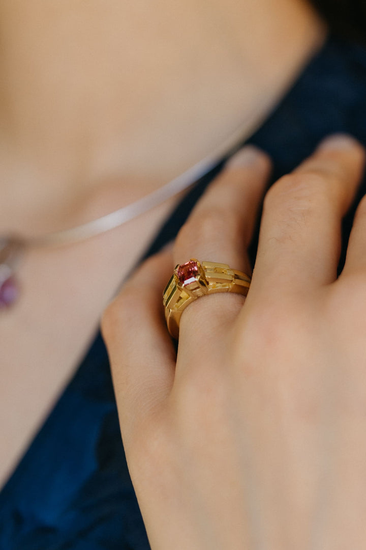 Nahaufnahme von Hand und schimmerndem Le Pont Ring aus Gelbgold und pinkem Turmalin, getragen in Kombination mit einer Halskette im Hintergrund