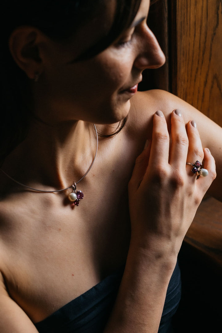Nahaufnahme eines eleganten Mon Amour Anhängers und Rings aus Weißgold mit Perle und violetten Edelsteinen am Hals und an der Hand einer Frau.