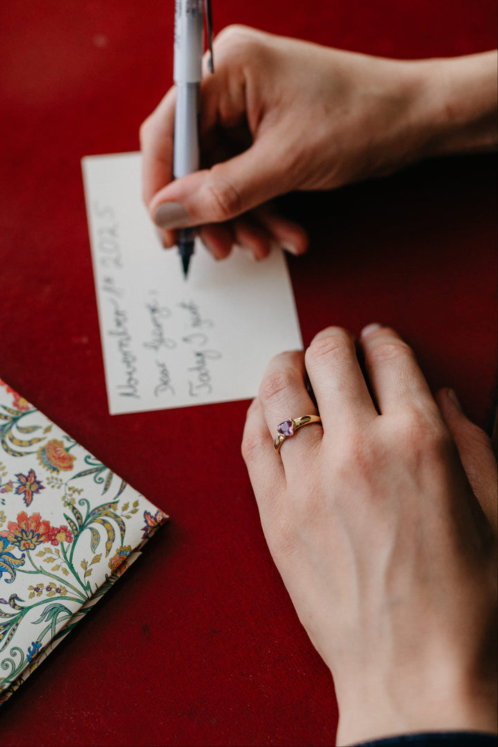 Mon Cœur Ring aus Gelbgold mit herzförmigem Edelstein auf einer schreibenden Hand, daneben ein florales Notizbuch.