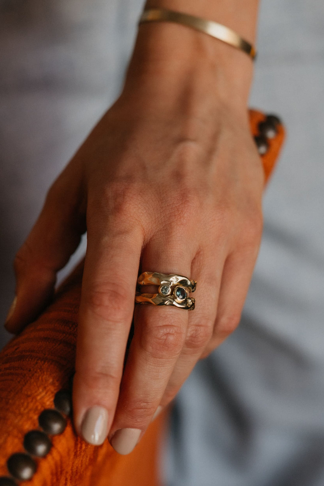 Mystery Ring aus Gold mit blauem Edelstein, am Finger einer Person, die eine Stuhllehne hält.
