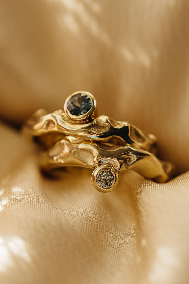 Goldener Mystery Ring mit organischer, geschwungener Oberfläche und rund gefasstem Turmalin, elegant auf goldenem Stoff fotografiert.