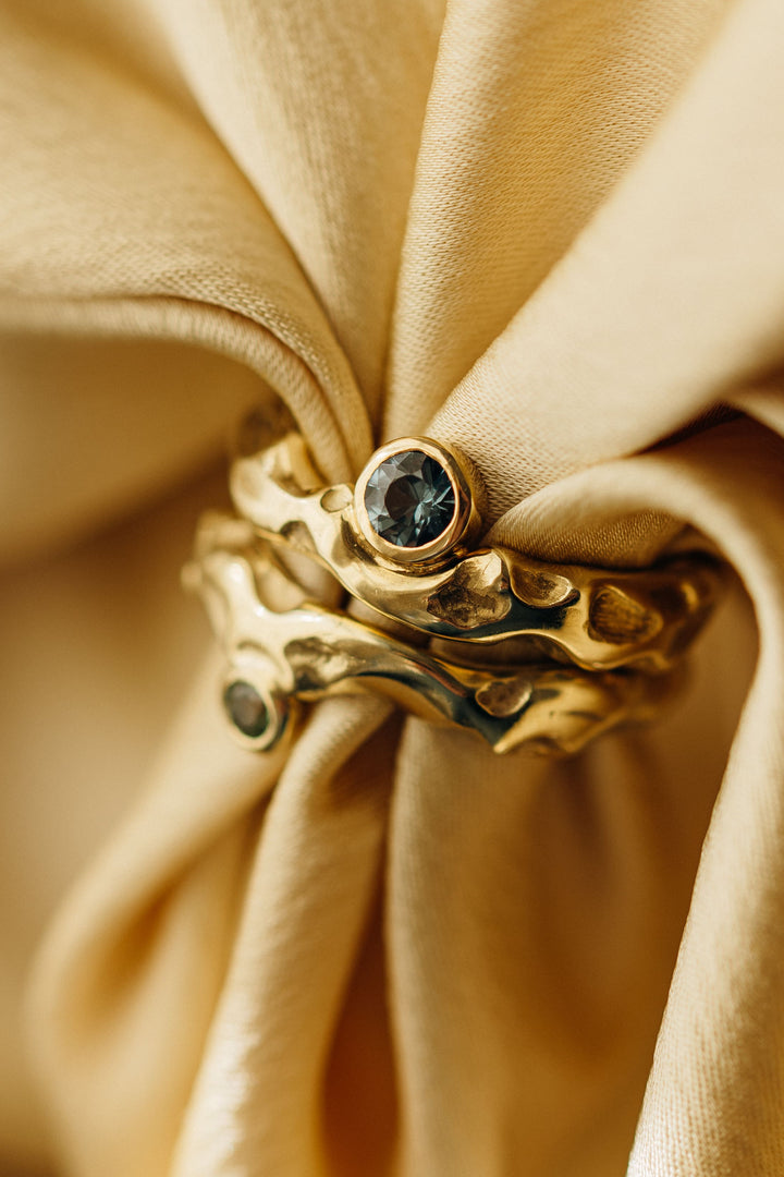 Mystery Single Ring aus Gold mit organischer Struktur und rund gefasstem Turmalin, fotografiert auf goldenem Stoff.