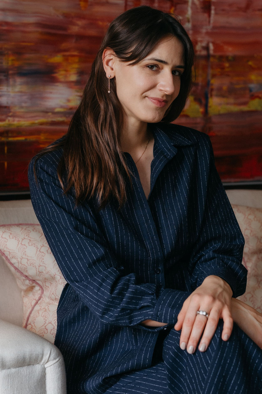 Portraitaufnahme einer Person, die Valentina-Halskette, Ohrringe und Ring trägt und auf einem Sofa sitzt und in die Kamera guckt.